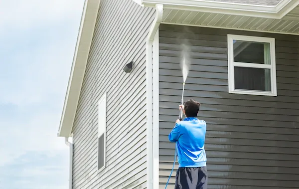 Person in a blue shirt using a pressure washer wand to clean gray vinyl siding on a two-story house